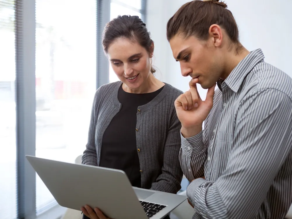 One to one training - a trainer assists a woman holding an open laptop, they are in an office location and both are looking at the laptop screen